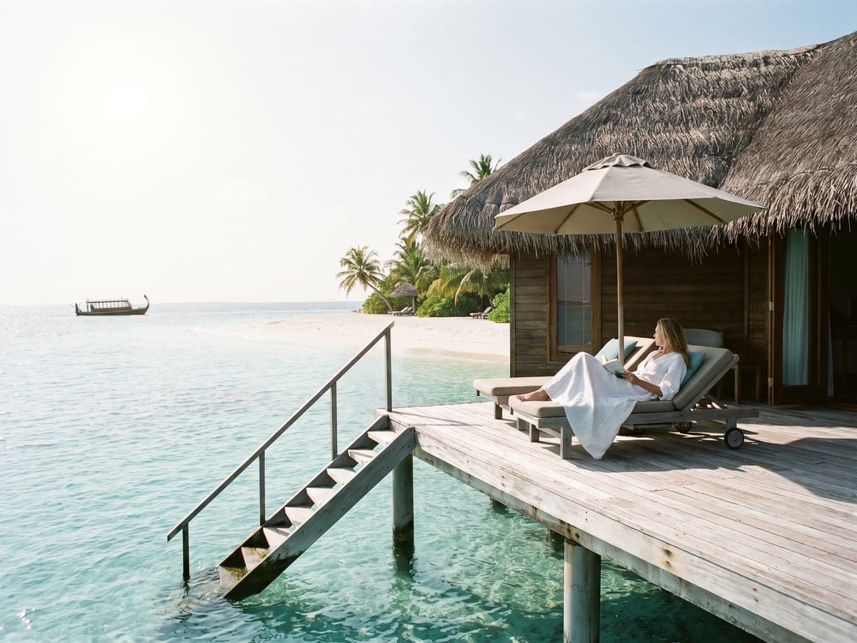 A thatched overwater villa above clear turquoise water, a woman reading on a daybed on its wooden deck
