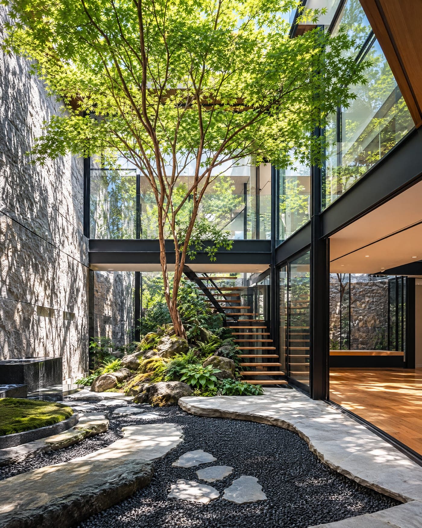 The courtyard garden of a glass and steel residence: a Japanese maple over mossy boulders, black gravel and stepping stones beside a stone wall